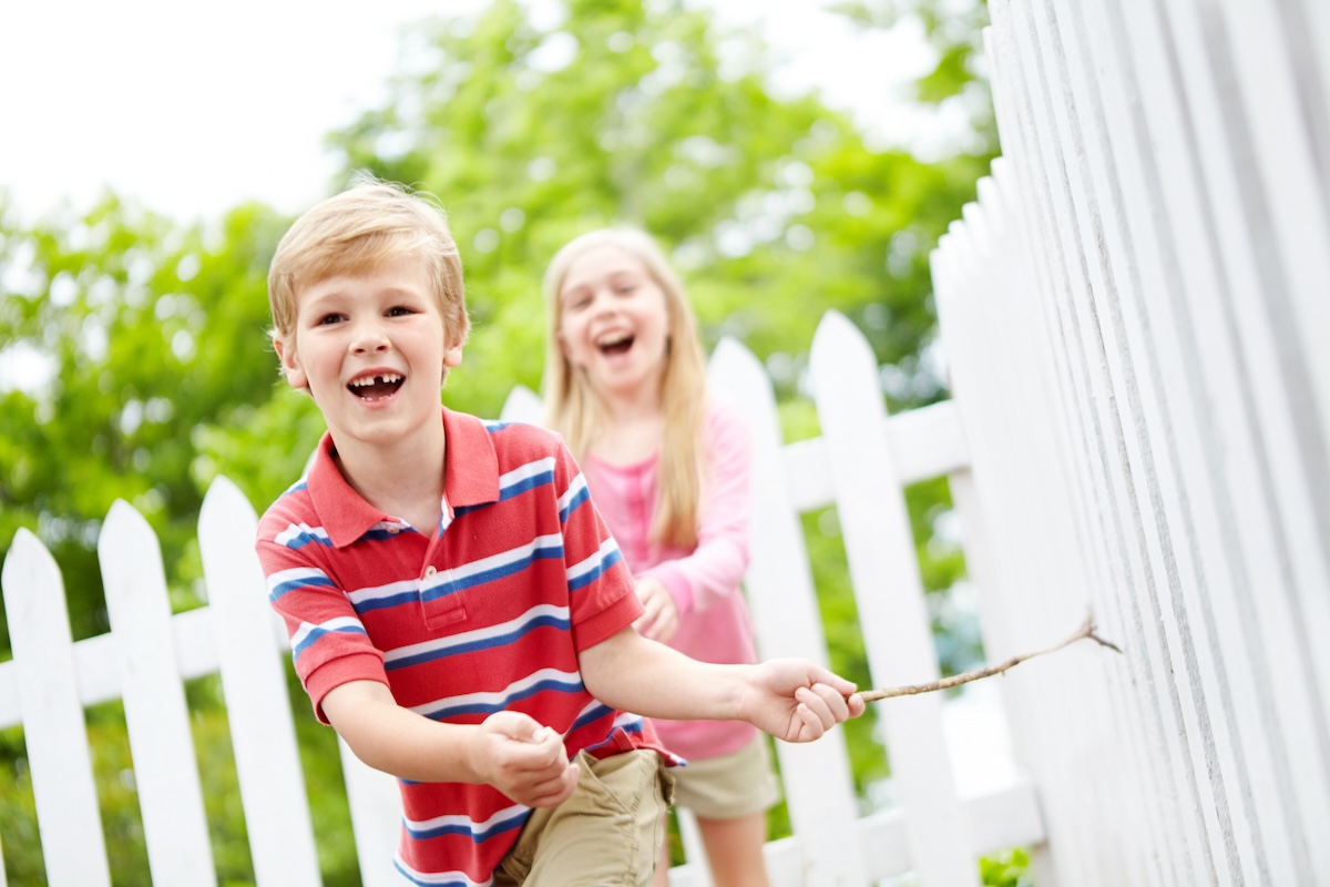 A cute young boy running a stick along a white vinyl picket fence while his sister chases him